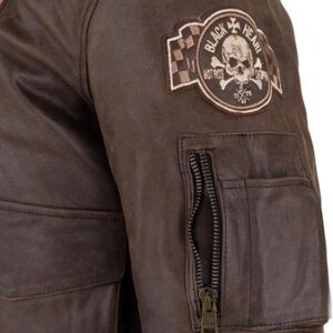 Op de afbeelding: Bruine leren jas met een schedel- en gekruiste botten-patch met de tekst "Black Heart Hot Rods". De jas heeft een zak met ritssluiting op de mouw en twee voorzakken. Het leer heeft een distressed look.