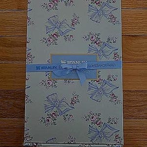 K&ouml;nnte beinhalten: Eine rechteckige Schachtel mit einem Blumen- und Schleifenmuster in Hellblau, Rosa und Wei&szlig;. Ein hellblaues Band mit dem Text "STANLEY LOVESHACKFANCY" umschlie&szlig;t die Schachtel. Die Schachtel hat ein hellblaues Etikett mit dem Text "STANLEY".