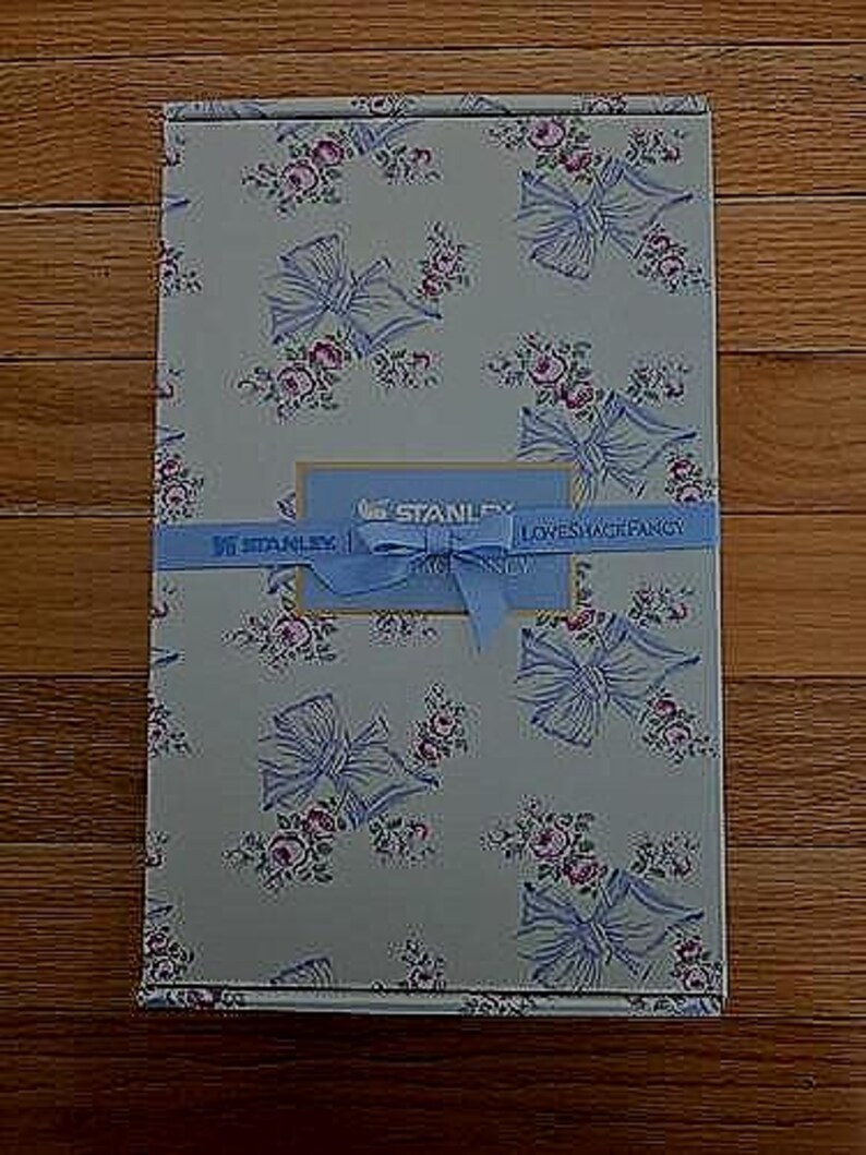 K&ouml;nnte beinhalten: Eine rechteckige Schachtel mit einem Blumen- und Schleifenmuster in Hellblau, Rosa und Wei&szlig;. Ein hellblaues Band mit dem Text "STANLEY LOVESHACKFANCY" umschlie&szlig;t die Schachtel. Die Schachtel hat ein hellblaues Etikett mit dem Text "STANLEY".