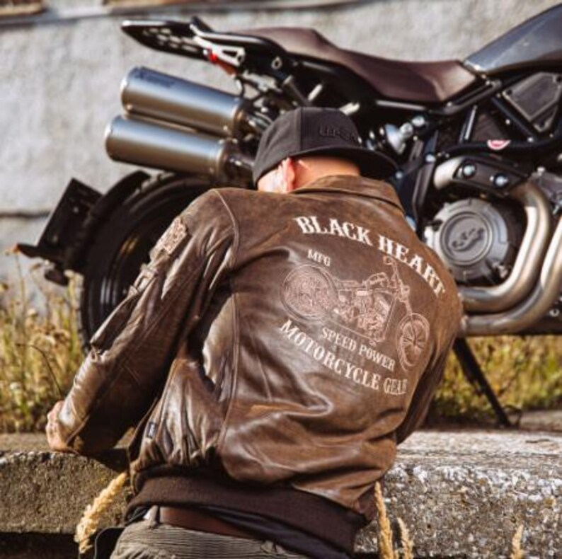 Op de afbeelding: Bruine leren motorjas met de tekst "BLACK HEART MOTORCYCLE GEAR" en een motorfietsafbeelding op de rug. De jas heeft een bomberstijl met een geribbelde zoom en manchetten. Een zwarte baseballcap wordt gedragen.