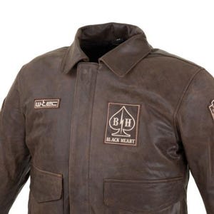 Op de afbeelding: Bruine leren jas met een kraag en twee voorzakken. De jas heeft patches met de tekst "wtec" en "BLACK HEART" met een schoppensymbool. De jas heeft een vintage uitstraling.