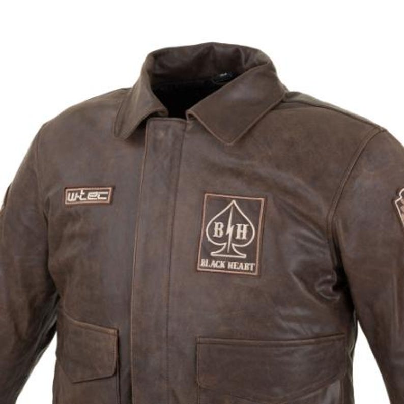 Op de afbeelding: Bruine leren jas met een kraag en twee voorzakken. De jas heeft patches met de tekst "wtec" en "BLACK HEART" met een schoppensymbool. De jas heeft een vintage uitstraling.