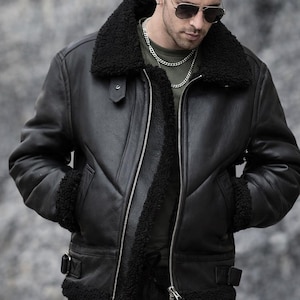 May include: Black leather jacket with a shearling collar and lining. The jacket has a zip-up front, pockets, and buckle details. The model is wearing sunglasses and a silver chain. A classic outerwear piece.