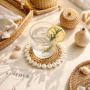 May include: A round woven rattan coaster decorated with white cowrie shells around the edge, supporting a clear ribbed glass filled with ice, a lemon slice, and a sprig of rosemary, positioned near a Kinfolk book and various woven decor items.