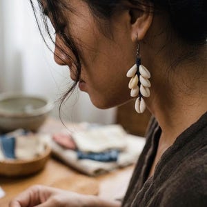 May include: Earrings featuring a cluster of cream-colored seashells suspended from a dark blue cord. The earrings are attached to a silver-colored hook. The earrings are a natural, beach-inspired accessory.