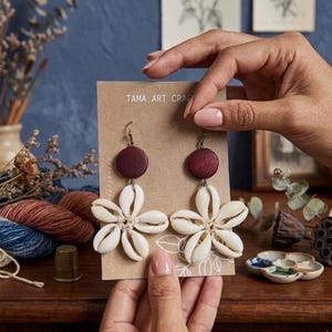Op de afbeelding: Handgemaakte oorbellen met een donkerrode houten schijf, een witte schelpenbloem en zilverkleurige haken. De oorbellen worden gepresenteerd op een bruine kaart met de tekst "TAMA ART CRAFTS".