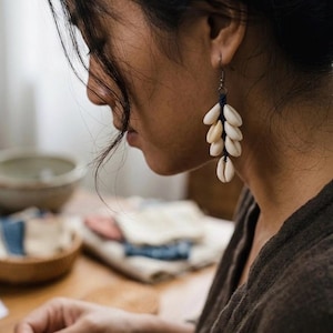 Op de afbeelding: Oorbellen met een cluster van crèmekleurige schelpen die aan een donkerblauw koord hangen. De oorbellen zijn bevestigd aan een zilverkleurige haak. Een natuurlijk, op het strand geïnspireerd accessoire.