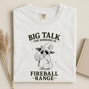 Pode incluir: Camiseta branca com um gr&aacute;fico de um guaxinim usando um chap&eacute;u e capa de mago, segurando um dado em chamas. O texto diz "BIG TALK for SOMEONE IN FIREBALL RANGE."