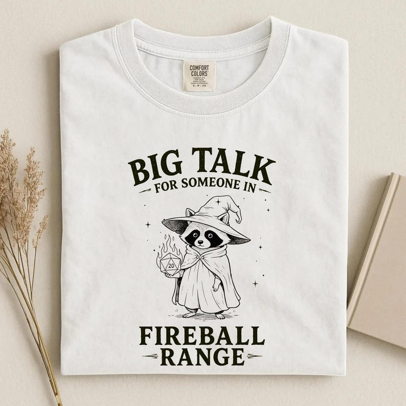 Pode incluir: Camiseta branca com um gr&aacute;fico de um guaxinim usando um chap&eacute;u e capa de mago, segurando um dado em chamas. O texto diz "BIG TALK for SOMEONE IN FIREBALL RANGE."