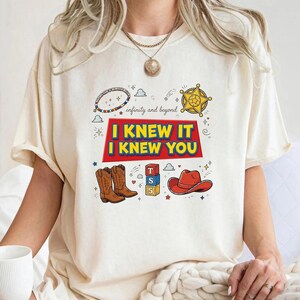 K&ouml;nnte beinhalten: Ein cremefarbenes T-Shirt mit einem Grafikdruck, der den Text "I KNEW IT I KNEW YOU" und "infinity and beyond" enth&auml;lt. Das Design zeigt Illustrationen eines Sheriffsterns, Cowboystiefeln, eines roten Cowboyhuts und Spielzeugw&uuml;rfeln.