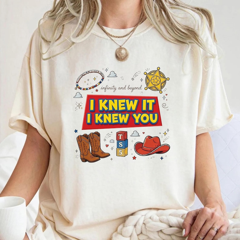K&ouml;nnte beinhalten: Ein cremefarbenes T-Shirt mit einem Grafikdruck, der den Text "I KNEW IT I KNEW YOU" und "infinity and beyond" enth&auml;lt. Das Design zeigt Illustrationen eines Sheriffsterns, Cowboystiefeln, eines roten Cowboyhuts und Spielzeugw&uuml;rfeln.
