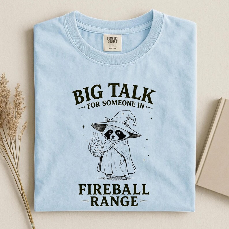 Pode incluir: Camiseta azul clara com o texto "BIG TALK FOR SOMEONE IN FIREBALL RANGE". A camiseta apresenta uma ilustra&ccedil;&atilde;o em preto e branco de um guaxinim usando um chap&eacute;u e capa de mago, segurando um dado em chamas.