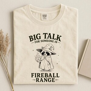 Pode incluir: Camiseta creme com o texto "BIG TALK FOR SOMEONE IN FIREBALL RANGE". Um guaxinim de desenho animado usando um chap&eacute;u e capa de mago segura um dado em chamas. A camisa tem uma etiqueta Comfort Colors.