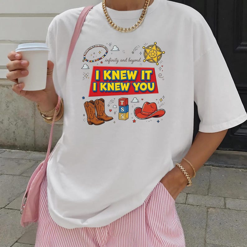 K&ouml;nnte beinhalten: Ein wei&szlig;es Kurzarm-T-Shirt mit einem Grafikdruck, der den Text "I KNEW IT I KNEW YOU" in gelben Buchstaben auf rotem Grund zeigt, erg&auml;nzt durch Illustrationen von braunen Cowboystiefeln, einem roten Cowboyhut, einem Sternabzeichen und Baukl&ouml;tzen.