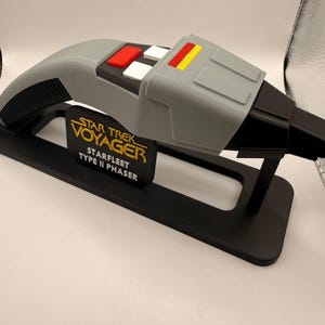 May include: A grey Star Trek Voyager Type II Phaser replica. The prop has red, white, and yellow accents on top, with the Starfleet logo on a black stand. The phaser is displayed on a black stand.