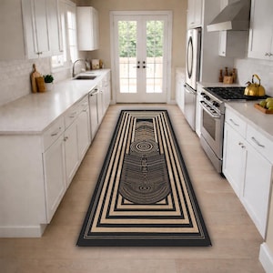 May include: A rectangular kitchen rug with a geometric design. The rug features a black and tan color scheme with a central abstract pattern and a series of concentric rectangles. The rug is placed in a kitchen with white cabinets and appliances.