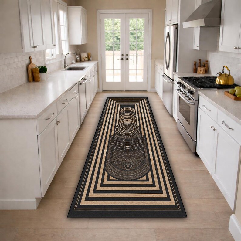 May include: A rectangular kitchen rug with a geometric design. The rug features a black and tan color scheme with a central abstract pattern and a series of concentric rectangles. The rug is placed in a kitchen with white cabinets and appliances.