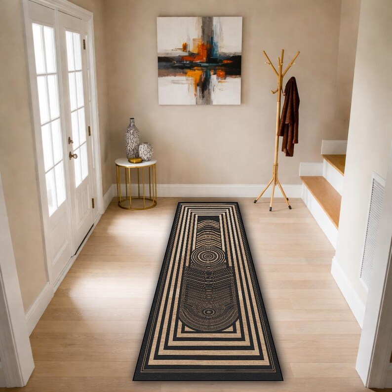May include: A rectangular black and beige geometric patterned rug. The rug features a central design with concentric circles and rectangular borders. The rug is placed in a hallway with light-colored wooden flooring and white doors.