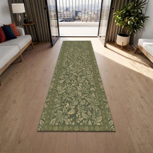 Op de afbeelding: Een lange, rechthoekige groene vloerkleed met een gedetailleerd bloemmotief. Het vloerkleed heeft een rand met een herhalend ontwerp en ligt op een houten vloer. Het ontwerp van het vloerkleed omvat bladeren en bloemen in groen- en beigetinten.