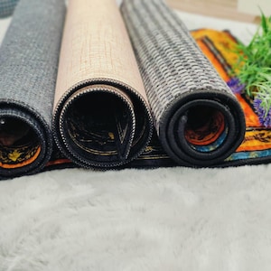May include: Three rolled-up decorative mats in shades of gray, beige, and patterned designs. The mats are rolled up, revealing their underside with intricate patterns and colors. The mats are placed on a white, fluffy surface.