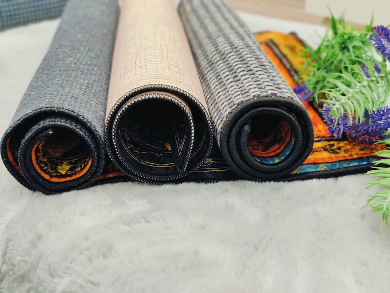 May include: Three rolled-up decorative mats in shades of gray, beige, and patterned designs. The mats are rolled up, revealing their underside with intricate patterns and colors. The mats are placed on a white, fluffy surface.
