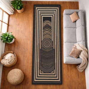 May include: A rectangular rug with a black and beige geometric design. The rug features a central pattern of concentric circles and curved lines within a black border. The rug is placed on a wooden floor, with a sofa and plants visible.