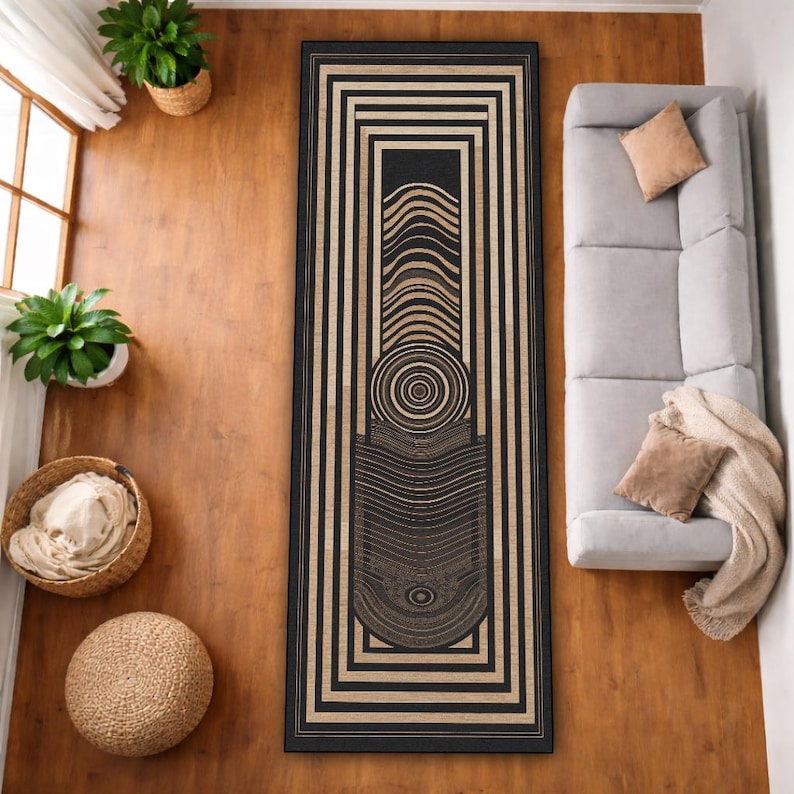 May include: A rectangular rug with a black and beige geometric design. The rug features a central pattern of concentric circles and curved lines within a black border. The rug is placed on a wooden floor, with a sofa and plants visible.