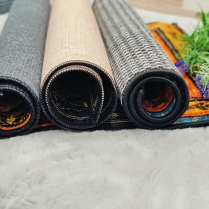 Puede incluir: Alfombras decorativas enrolladas en tonos grises, beige y dise&ntilde;os estampados. Los bordes son oscuros con un borde contrastante. Las alfombras se exhiben sobre una superficie blanca y esponjosa, con vegetaci&oacute;n y flores moradas en el fondo.