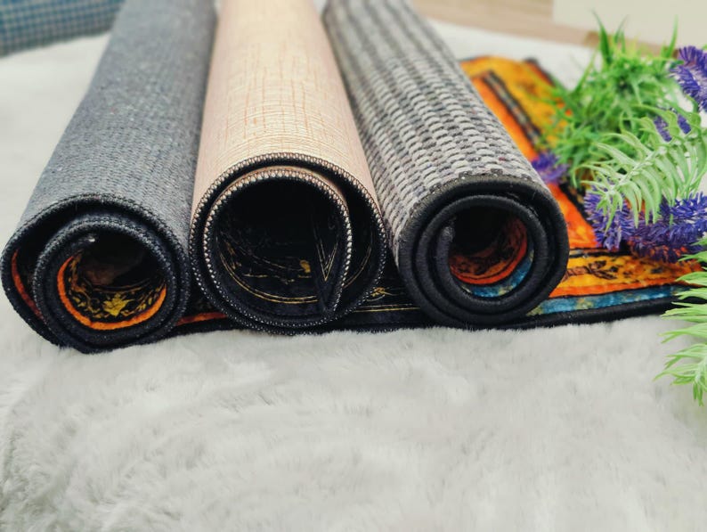 Puede incluir: Alfombras decorativas enrolladas en tonos grises, beige y dise&ntilde;os estampados. Los bordes son oscuros con un borde contrastante. Las alfombras se exhiben sobre una superficie blanca y esponjosa, con vegetaci&oacute;n y flores moradas en el fondo.