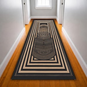 May include: A long, rectangular rug with a geometric design in black and tan. The rug features concentric circles and a rectangular pattern, creating a modern, abstract look. The rug is placed in a hallway with wooden floors.