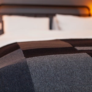 May include: A patchwork blanket in shades of grey, black, and brown covers a bed with white sheets. The blanket's design features large, rectangular blocks of colour, creating a geometric pattern. The headboard is a dark metal frame.