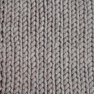 Puede incluir: Primer plano de un tejido de punto gris con un patr&oacute;n trenzado grueso. Las filas verticales del tejido crean una superficie texturizada. El color es un gris neutro, adecuado para varios art&iacute;culos de decoraci&oacute;n del hogar.