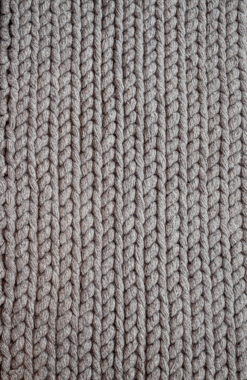 Puede incluir: Primer plano de un tejido de punto gris con un patr&oacute;n trenzado grueso. Las filas verticales del tejido crean una superficie texturizada. El color es un gris neutro, adecuado para varios art&iacute;culos de decoraci&oacute;n del hogar.