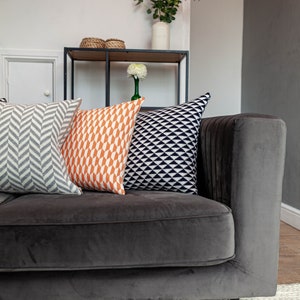 Puede incluir: Un sof&aacute; de terciopelo gris oscuro con tres cojines decorativos. Los cojines presentan patrones geom&eacute;tricos en gris, naranja y negro. Se ve una estanter&iacute;a con decoraci&oacute;n y un jarr&oacute;n con vegetaci&oacute;n.