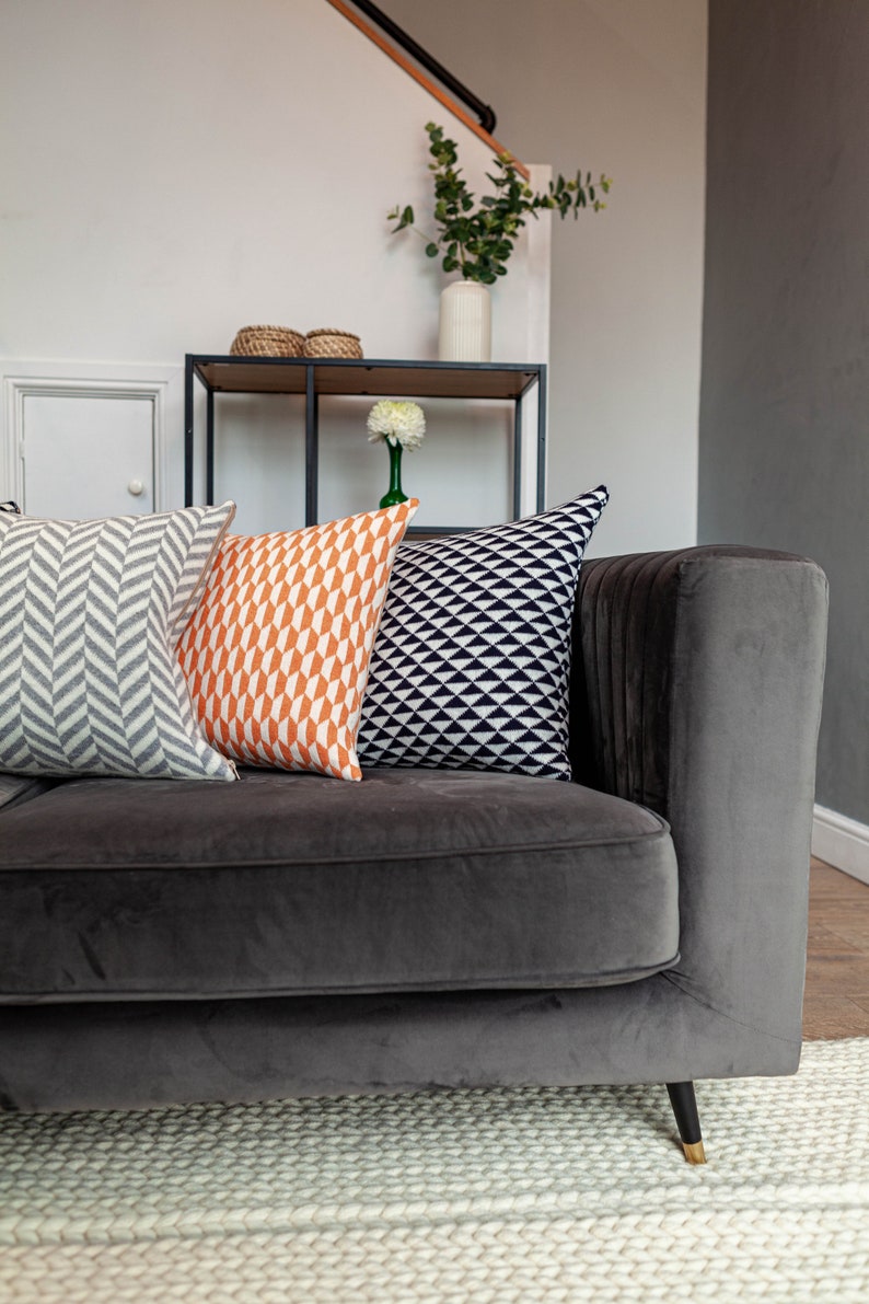 Puede incluir: Un sof&aacute; de terciopelo gris oscuro con tres cojines decorativos. Los cojines presentan patrones geom&eacute;tricos en gris, naranja y negro. Se ve una estanter&iacute;a con decoraci&oacute;n y un jarr&oacute;n con vegetaci&oacute;n.