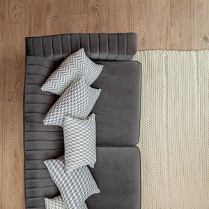 Puede incluir: Vista a&eacute;rea de una sala de estar moderna. Un sof&aacute; gris oscuro est&aacute; adornado con varias almohadas decorativas con patrones geom&eacute;tricos en gris y blanco. Una alfombra de punto grueso de color crema se encuentra junto al sof&aacute; sobre un piso de madera.