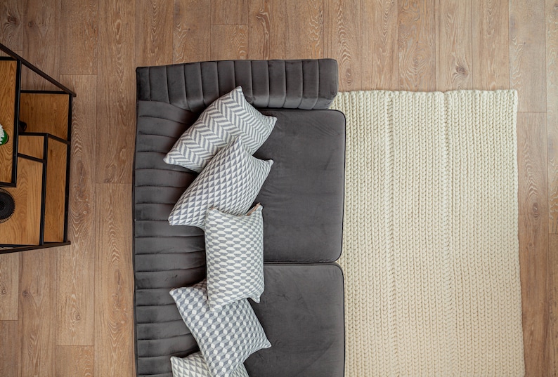 Puede incluir: Vista a&eacute;rea de una sala de estar moderna. Un sof&aacute; gris oscuro est&aacute; adornado con varias almohadas decorativas con patrones geom&eacute;tricos en gris y blanco. Una alfombra de punto grueso de color crema se encuentra junto al sof&aacute; sobre un piso de madera.