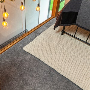 Puede incluir: Una alfombra de punto grueso de color crema sobre una superficie alfombrada gris. La alfombra es rectangular y parece estar tejida a mano. En el fondo se ven una estructura de cama de metal negro y una colcha de retazos. Luces decorativas colgando.