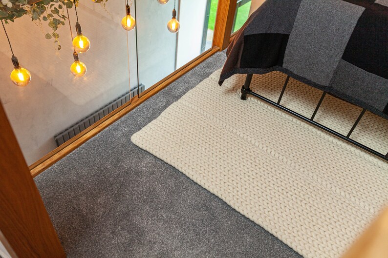 Puede incluir: Una alfombra de punto grueso de color crema sobre una superficie alfombrada gris. La alfombra es rectangular y parece estar tejida a mano. En el fondo se ven una estructura de cama de metal negro y una colcha de retazos. Luces decorativas colgando.