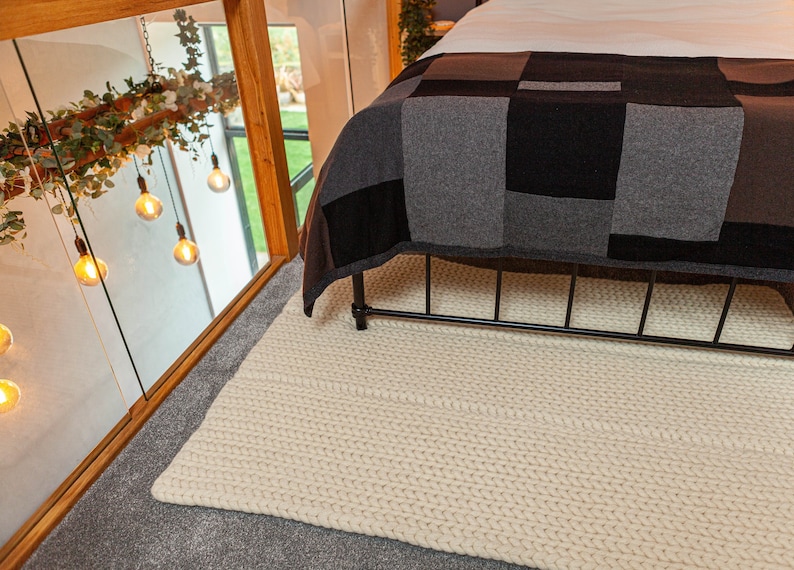 Puede incluir: Una alfombra de punto grueso de color crema se coloca sobre un suelo alfombrado gris, cerca de una cama con un marco de metal negro. La cama est&aacute; cubierta con un edred&oacute;n de retazos en tonos grises y negros. Las bombillas decorativas cuelgan del techo.