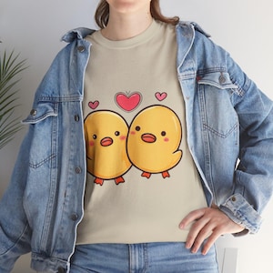 Op de afbeelding: Beige T-shirt met een cartoonontwerp van twee gele kuikens met oranje snavels en poten en roze wangen. Een rood hart en twee kleinere roze harten staan boven de kuikens. De persoon draagt een blauw spijkerjasje.