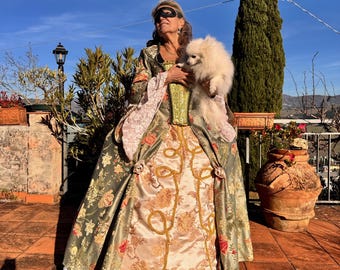 May include: A woman in a historical costume, holding a small white dog, stands on a terracotta patio. The dress is olive green with floral patterns, gold accents, and a corset-style bodice. She wears a black eye mask.