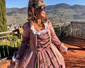 May include: A person wearing a pink and gold floral patterned dress with long sleeves and a white lace trim. The dress has a high waist and a full skirt. The person is also wearing a black eye mask and holding a fan.