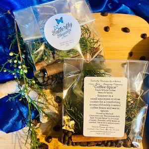 Op de afbeelding: Twee doorzichtige zakjes gevuld met koffiebonen, kruiden en specerijen. Eén zakje heeft een rond etiket met een vlinderlogo en de tekst "Butterfly Blessings". Het andere zakje heeft een etiket met de tekst "Coffee Spice".