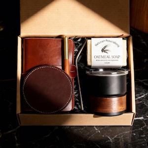 Könnte beinhalten: Geöffnete Geschenkbox mit einem braunen Lederjournal, Stift, Untersetzer und einem schwarz-braunen Becher. Enthält auch ein Stück Haferflocken-Seife und einen Metallstrohhalm. Die Artikel sind in der Box angeordnet.