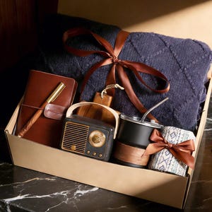 Könnte beinhalten: Eine Geschenkbox gefüllt mit einer marineblauen Decke, die mit einem braunen Band gebunden ist, einem braunen Lederjournal mit einem goldenen Stift, einem Radio im Vintage-Stil, einem schwarz-holzfarbenen Becher und einem gemusterten Tuch, das mit einem braunen Band gebunden ist.