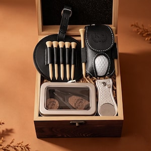 Könnte beinhalten: Ein Golf-Geschenkset in einer Holzkiste. Es enthält einen schwarzen Leder-Teehalter mit Golfball, Tees, ein Divot-Reparaturwerkzeug und eine Metalldose. Die Kiste ist dunkelbraun mit hellbraunem Innenraum.