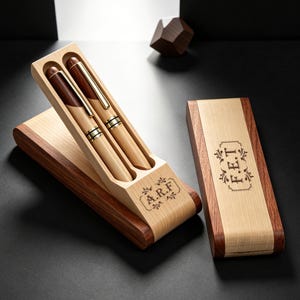 Könnte beinhalten: Zwei Holzstifte mit goldfarbenen Clips und Akzenten in einem Etui aus hellem und dunklem Holz mit den eingravierten Initialen A.R.F. Ein passender Etuideckel liegt daneben und zeigt die eingravierten Initialen F.E.T. innerhalb eines dekorativen Rahmens.