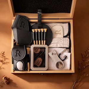 Könnte beinhalten: Eine Holzbox mit Golfzubehör. Das Set enthält einen schwarzen Golfballhalter aus Leder, einen Golfball, Tees, ein Divot-Reparaturwerkzeug, einen Golfhandschuh und ein Bürstenset. Das Zubehör ist in der Box angeordnet, mit dem Buchstaben "R" auf mehreren Artikeln.