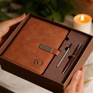 Puede incluir: Un juego de diario y bolígrafos de cuero marrón en una caja de regalo. El diario tiene un cierre plateado y el nombre "VIVIAN" con una corona de laurel. El juego incluye un bolígrafo plateado y un bolígrafo marrón. La caja es sostenida por manos.